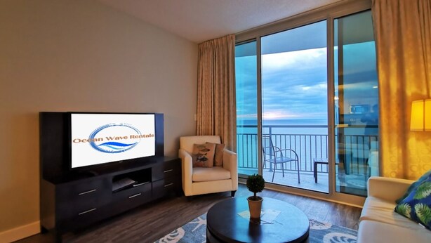 A living room with a television displaying 'Ocean Wave Rentals,' a sofa, a chair, a small table, and a balcony with a view.