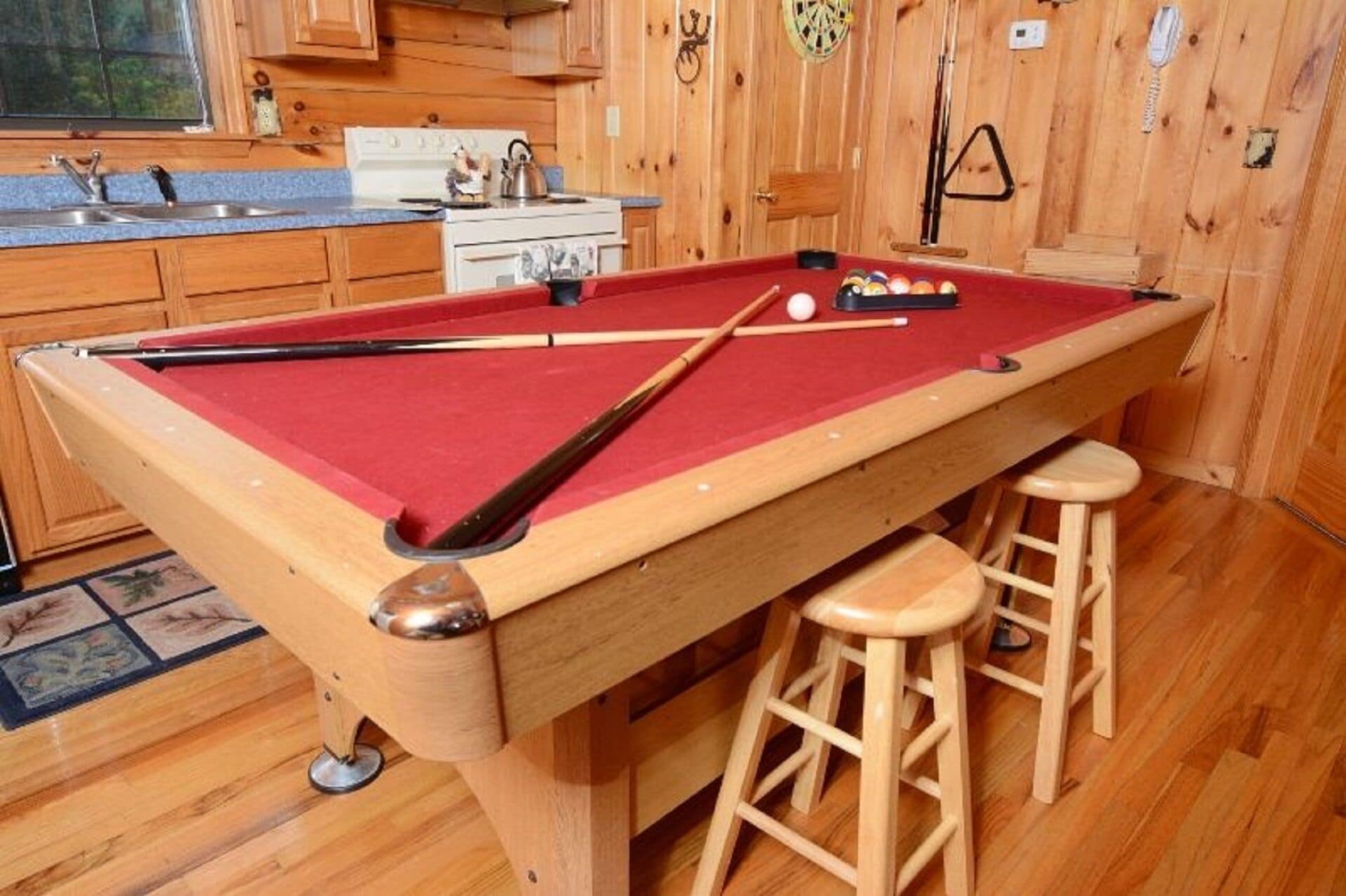 Una mesa de billar con taburetes, una estufa y armarios de madera en una habitación con suelo de madera y una ventana.