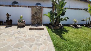 Une cour intérieure avec un chemin de pierre menant à un mur de pierre agrémenté d’un élément décoratif aquatique. On y trouve des plantes en pot et un petit espace jardin avec de l’herbe et diverses plantes.