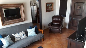 Un salon avec un canapé gris, des coussins à motifs, un fauteuil en cuir marron, un meuble en bois et un sol carrelé.