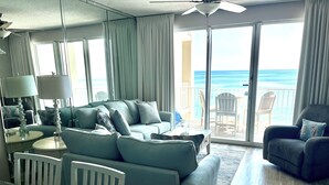 A modern living room with a large sofa, a glass coffee table, and a dining area with a glass table and chairs. The room has a view of the ocean through large sliding doors.