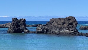 Una playa con aguas azules cristalinas, formaciones rocosas y vegetación verde en primer plano.