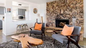 A living room with a stone wall, two grey armchairs with orange cushions, a wooden coffee table, and a fireplace. - Joshua Tree Getaway Private Desert Views (Yucca Valley)