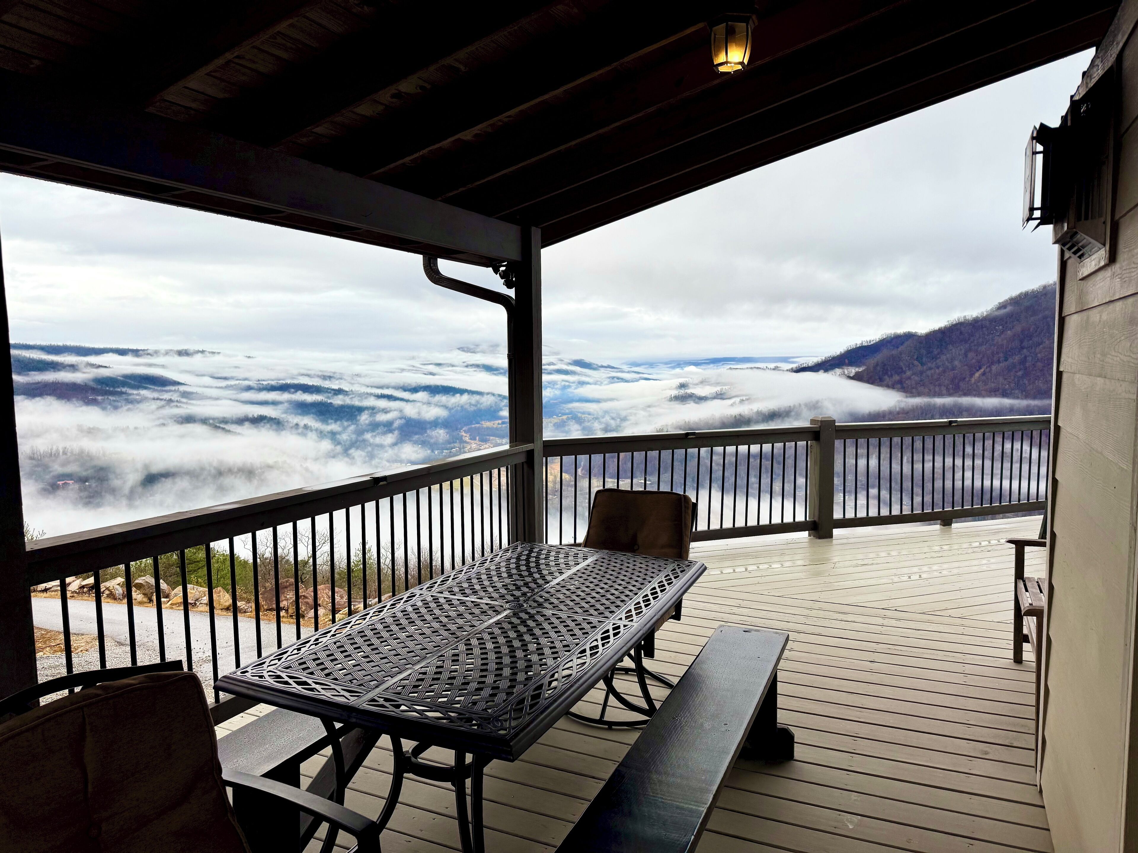 Un espacio de estar al aire libre techado con una mesa y sillas, con vistas a un paisaje montañoso y nubes abajo.