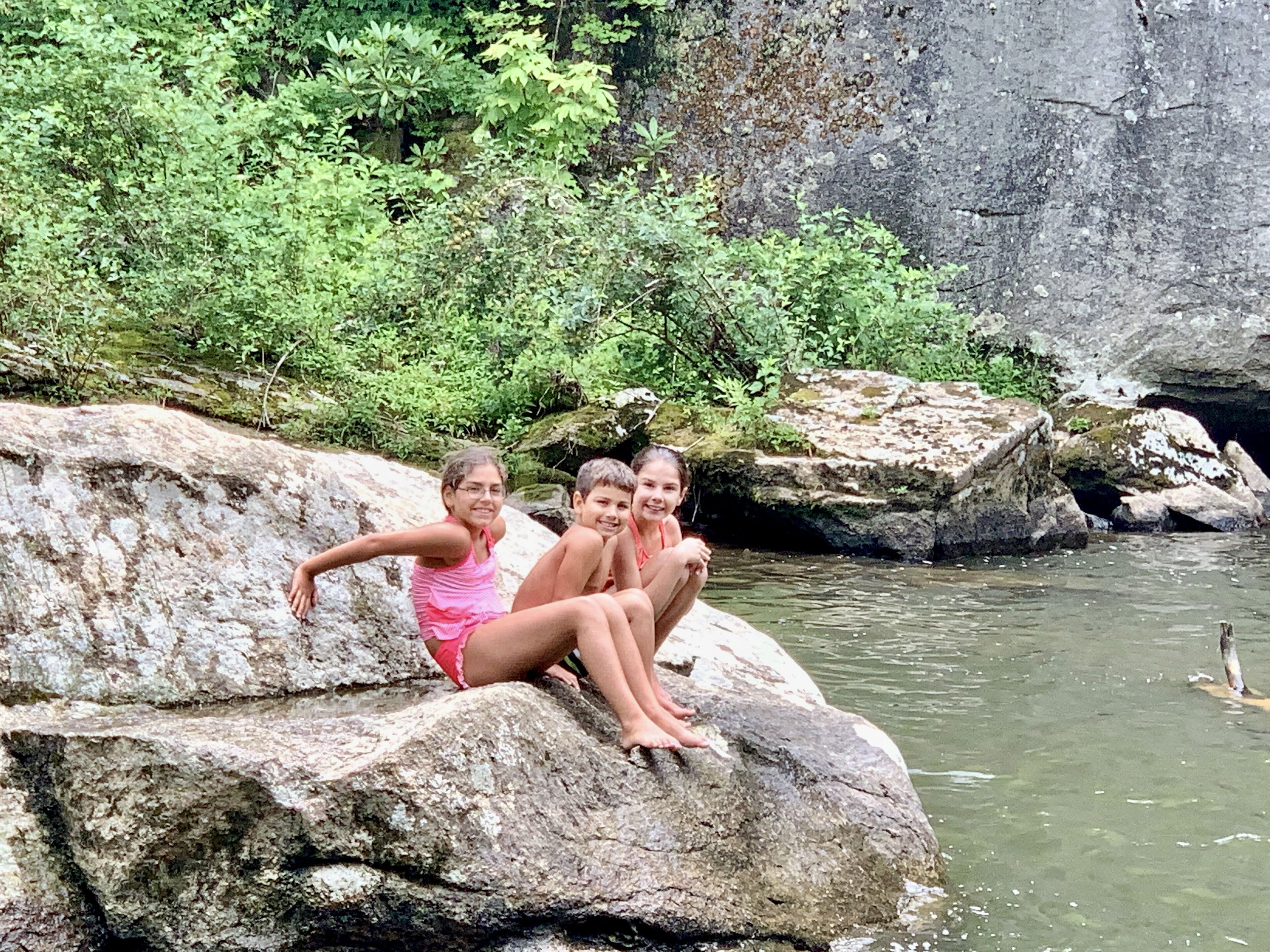 Three individuals sitting on a large rock by a body of water, surrounded by greenery and a rocky cliff.