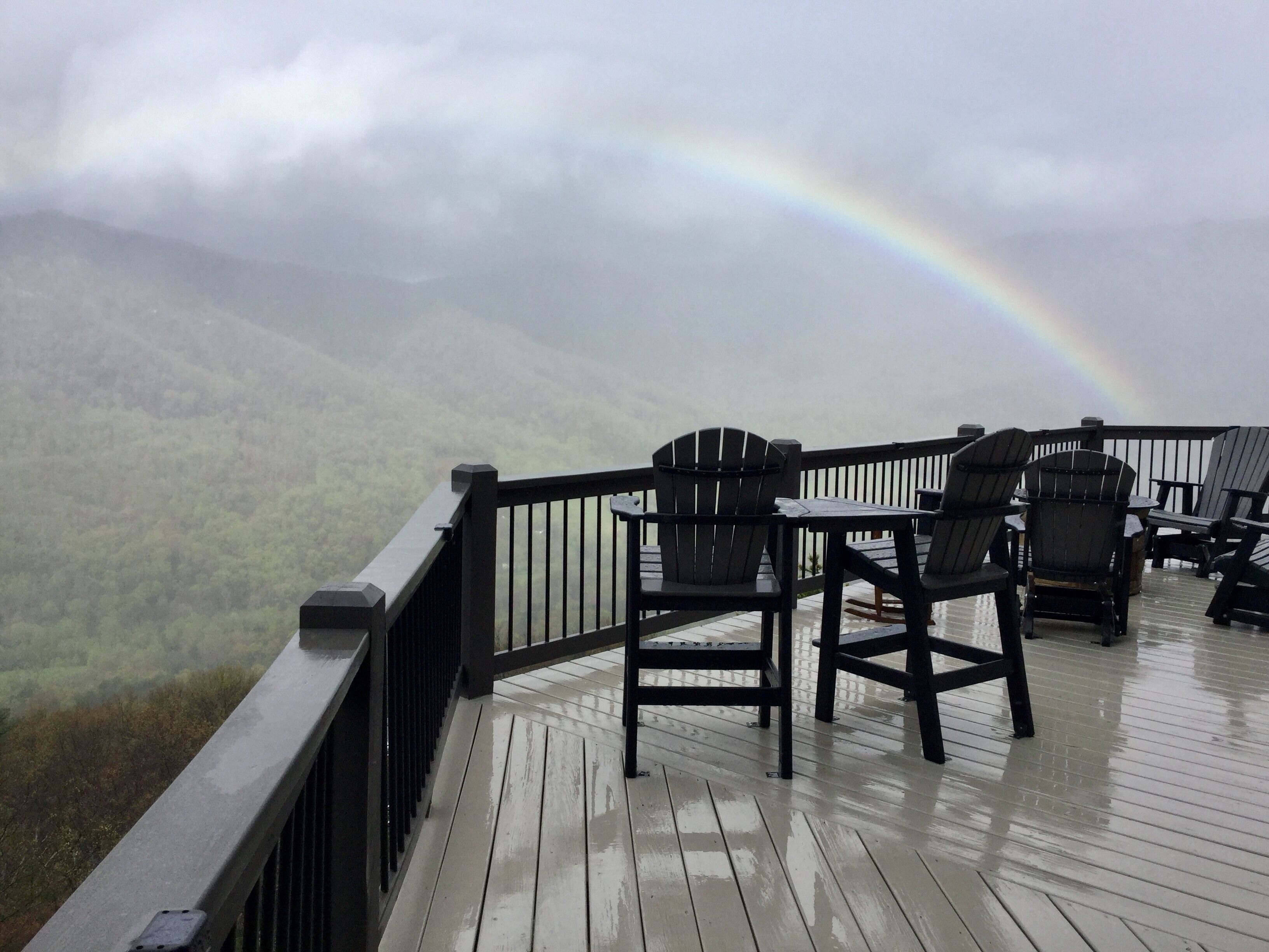 Una terraza con sillas Adirondack con vistas a un valle boscoso y un arcoíris.