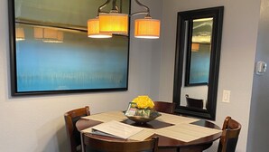 Un comedor con una mesa redonda, seis sillas de madera, una araña, un espejo y una obra de arte enmarcada en la pared.