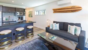 A modern living room with a grey sofa, three gold-striped stools, a glass coffee table, and a blue rug. - Villa 6 (Kihei)