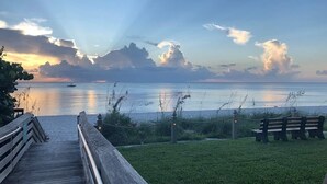 A wooden boardwalk leading to a grassy area with benches, overlooking a calm body of water and a sunset sky.