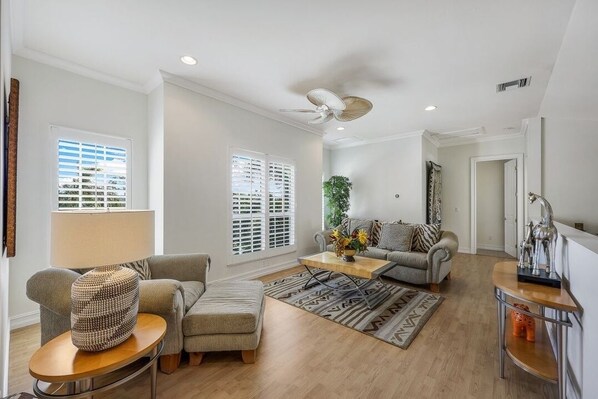 A spacious living room with a sofa, ottoman, and coffee table, a ceiling fan, and large windows with blinds.