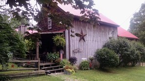 Une grange rustique avec un toit rouge, des marches en bois menant à un porche, ainsi que diverses plantes en pot et décorations disposées autour de l’entrée.