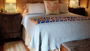 Une chambre confortable avec un grand lit, une tête de lit en bois et une couverture matelassée bleue. On y trouve deux tables de chevet avec des lampes, une commode en bois et un tapis tissé sur le plancher en bois.