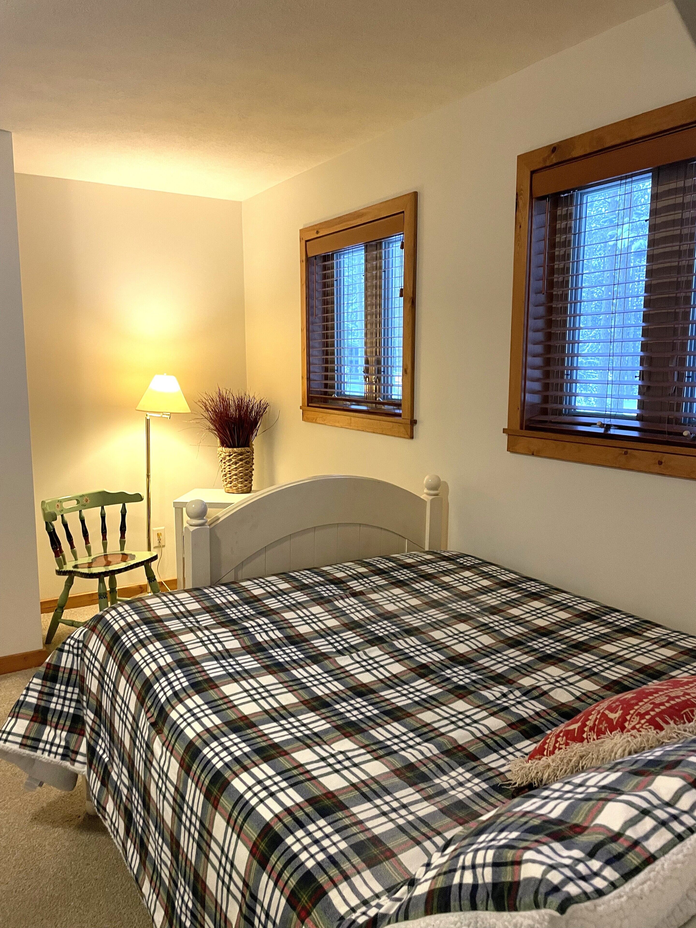 Quarto com cama, cadeira, luminária e duas janelas com persianas.