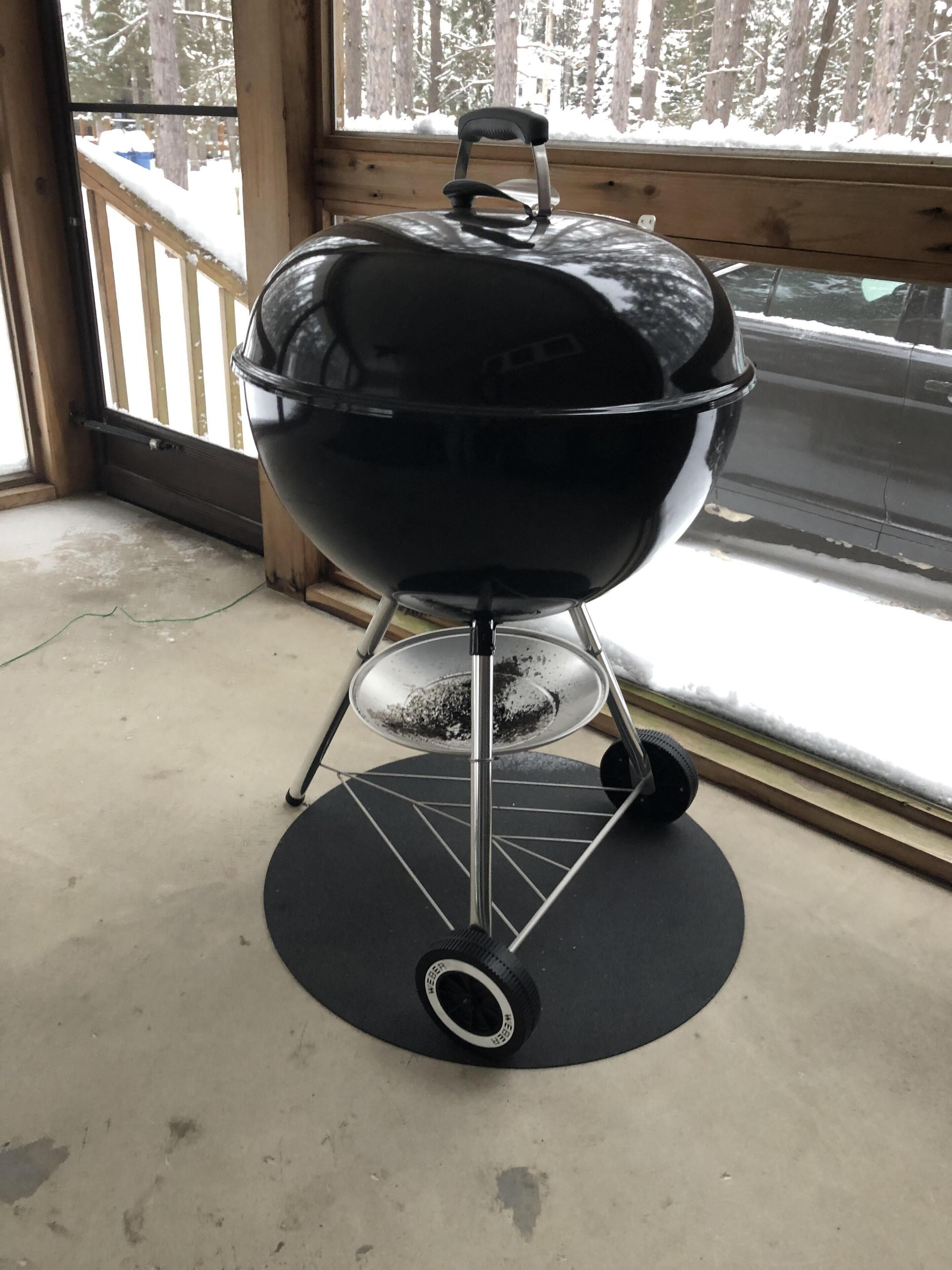 Un barbecue noir sur roulettes, doté d’une poignée, est placé sur un tapis noir à l’intérieur d’une pièce. On peut voir, à travers une fenêtre, une terrasse en bois.