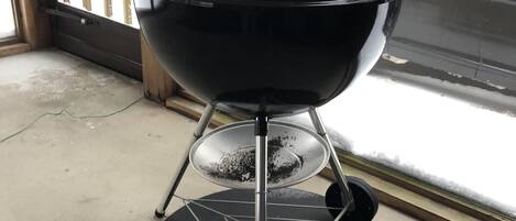 Un barbecue noir sur roulettes, doté d’une poignée, est placé sur un tapis noir à l’intérieur d’une pièce. On peut voir, à travers une fenêtre, une terrasse en bois.