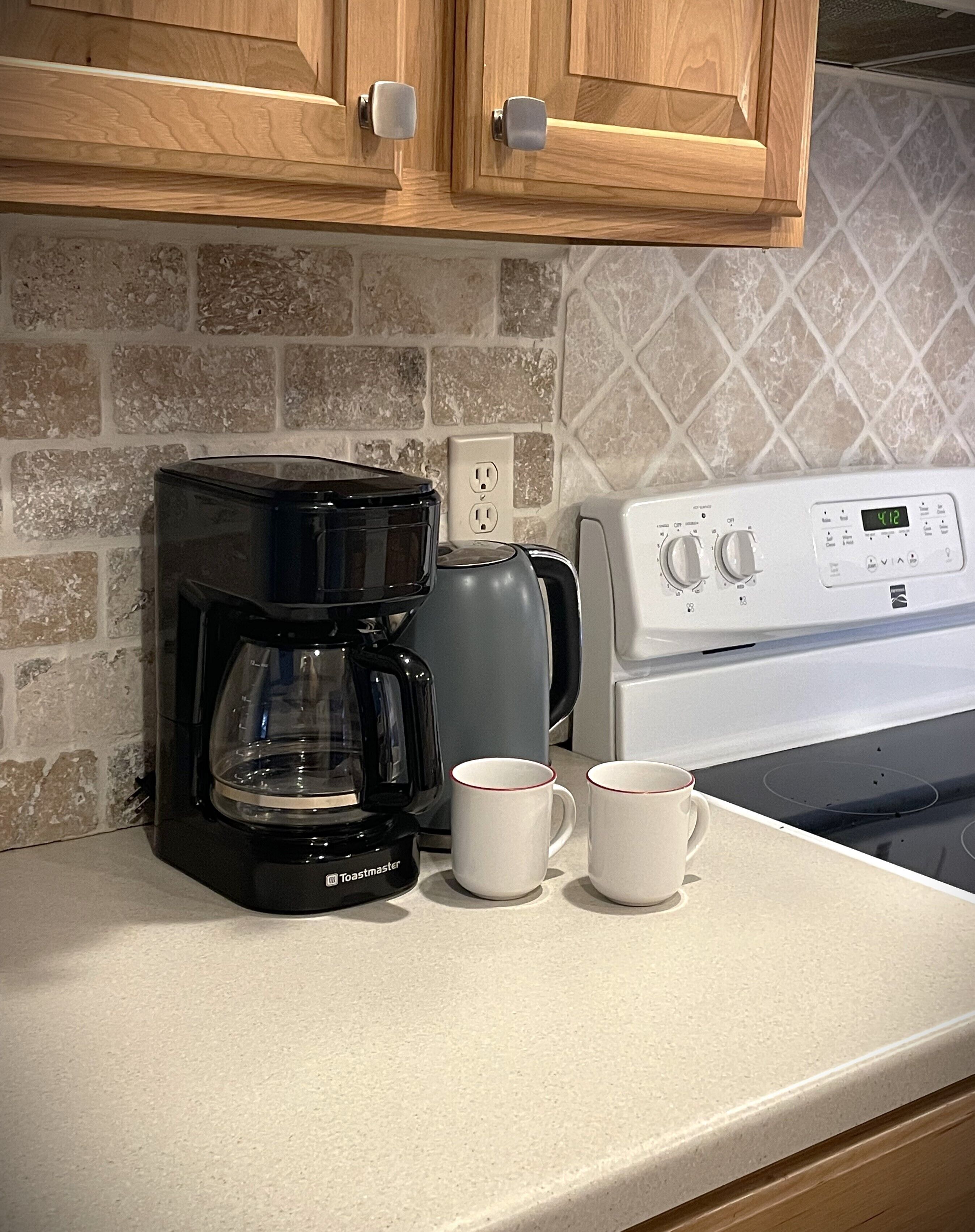 Uma cozinha com uma cafeteira Toastmaster preta, duas canecas brancas, uma chaleira elétrica cinza e um fogão branco.