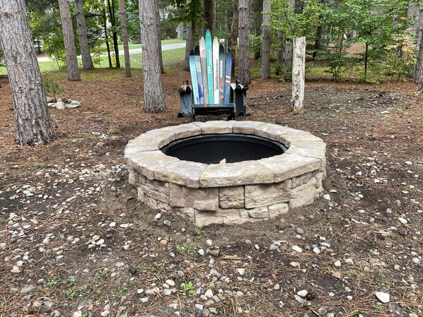 A stone fire pit surrounded by a forested area with trees and a metal chair with skis leaning against it. - Beautiful private cabin inside Crystal Mountain Resort (Thompsonville)