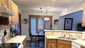 Una cocina con armarios de madera, una mesa de comedor con sillas, una araña y una ventana con persianas.