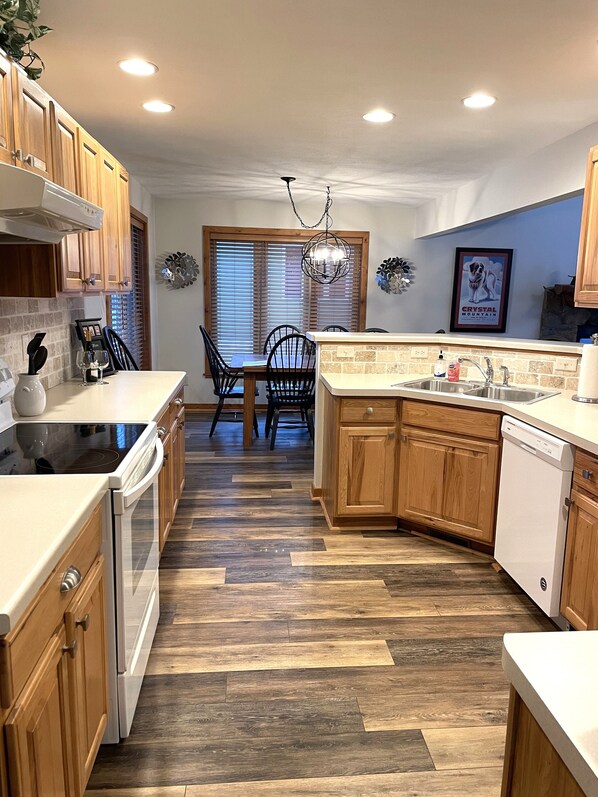 A kitchen with wooden cabinets, a dining table with chairs, a chandelier, and a window with blinds.