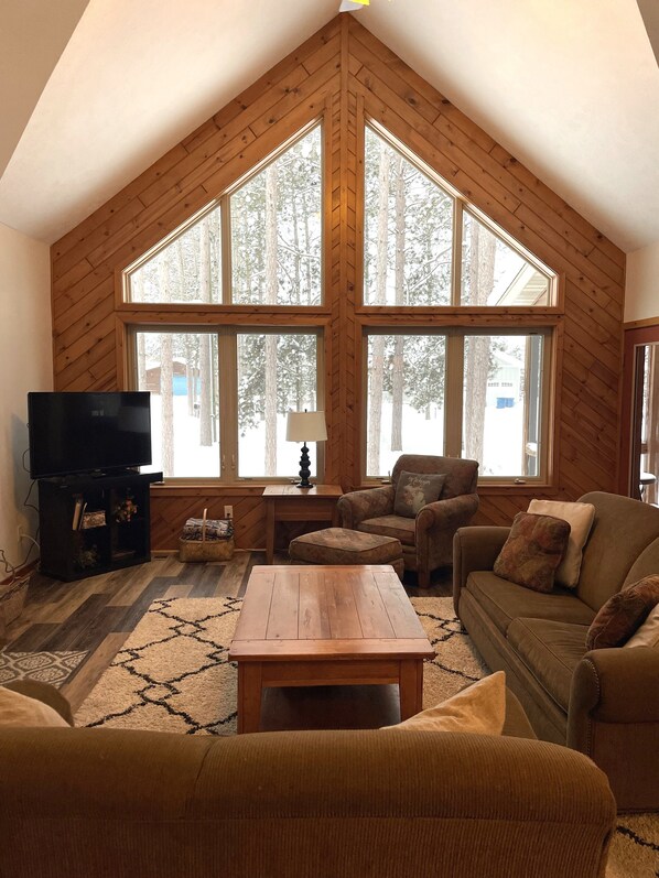 A cozy living room with a high ceiling, wooden walls, and large windows offering a view of trees.