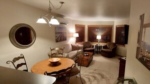 A cozy living room with a round wooden table, two armchairs, a sofa, and a TV. There are two lamps on side tables, a painting on the wall, and a window with blinds.
