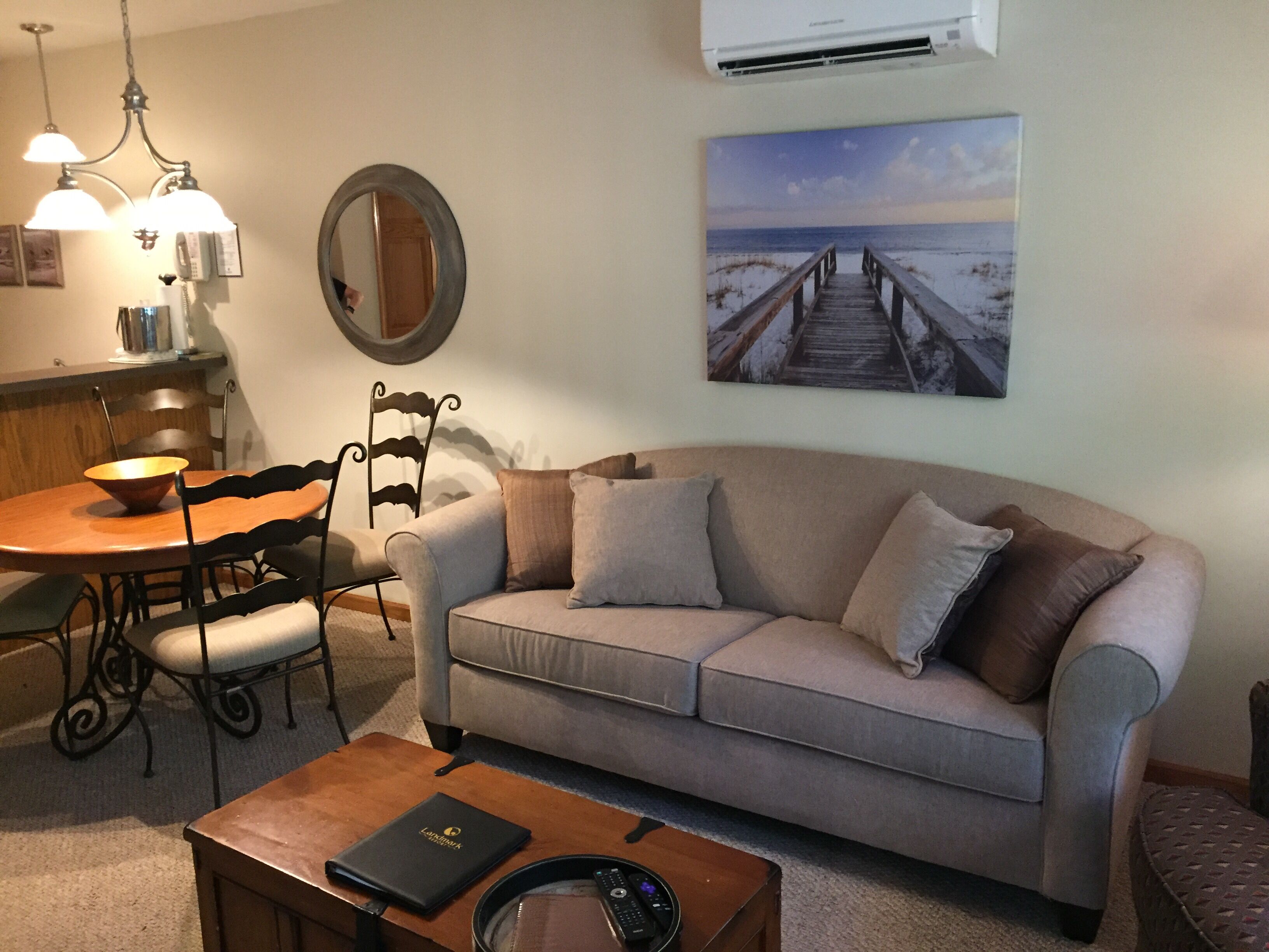 A living room with a sofa, a dining table with chairs, a coffee table, a mirror, and a painting of a beach scene.