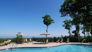 A pool area with lounge chairs and umbrellas, surrounded by trees and a clear view of the ocean.