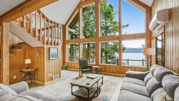 Un vaste salon doté d’une grande fenêtre donnant sur un lac, d’un escalier avec des poutres en bois et d’un coin salon confortable.