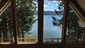 Une vue à travers une grande fenêtre sur une terrasse en bois aménagée avec du mobilier d’extérieur, donnant sur un lac paisible et des montagnes lointaines.