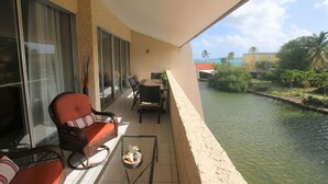 A balcony with wicker furniture, a glass table, and a view of a water body and greenery.