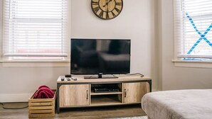 Une chambre à coucher avec un téléviseur posé sur un meuble en bois, une horloge au mur et une fenêtre avec des stores.