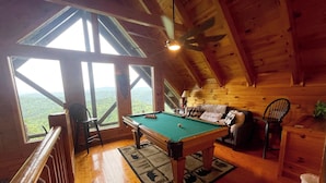 A wooden cabin with a pool table, a sofa, and a chair. There are large windows offering a view of the forest.