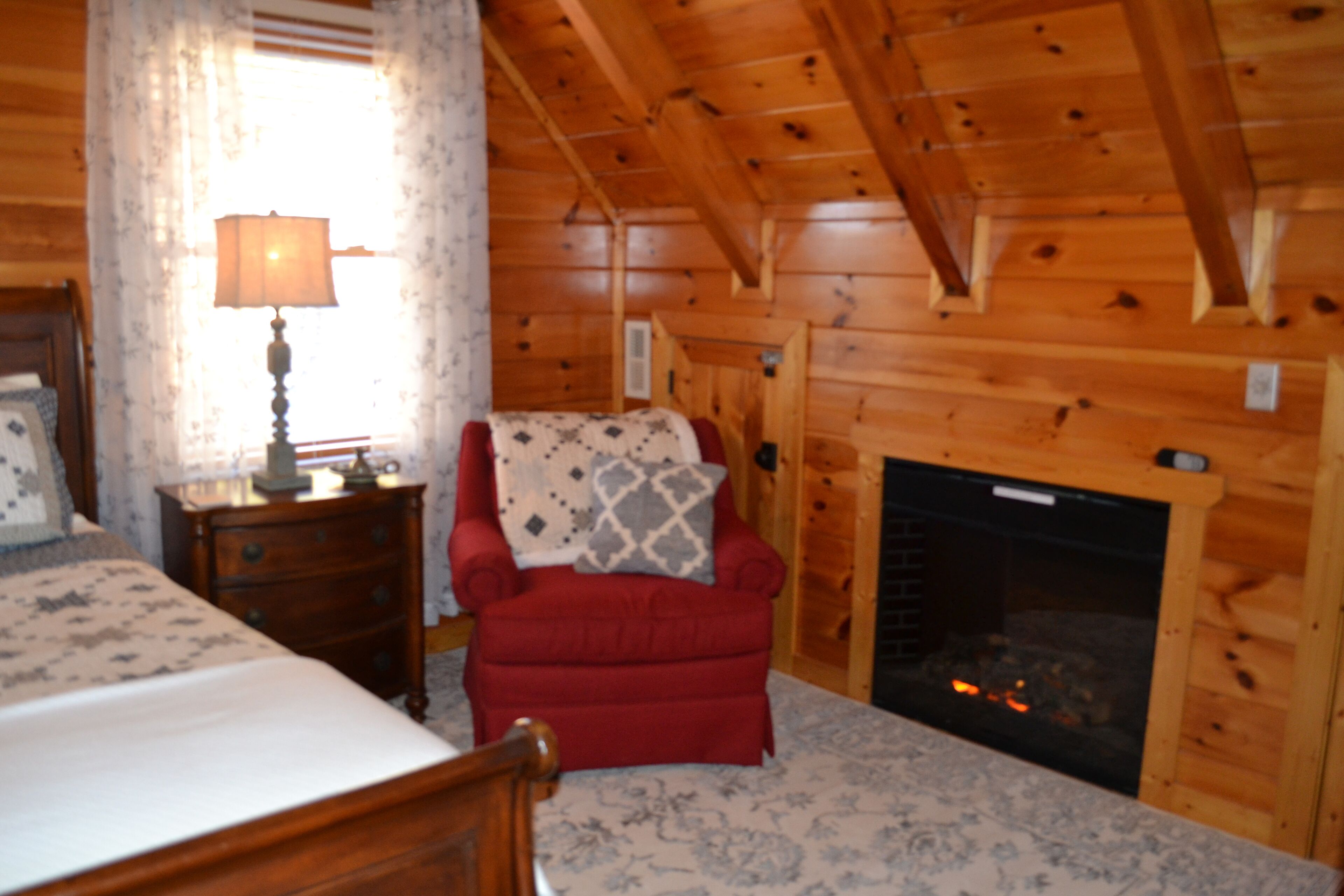 A wooden cabin interior with a bed, a red armchair, a fireplace, and a nightstand with a lamp.