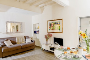 A living room with a brown sofa, a TV, a painting, and a dining area with a table set for breakfast. - A Few Steps From  Duomo Cathedral and SMN train station (Firenze)
