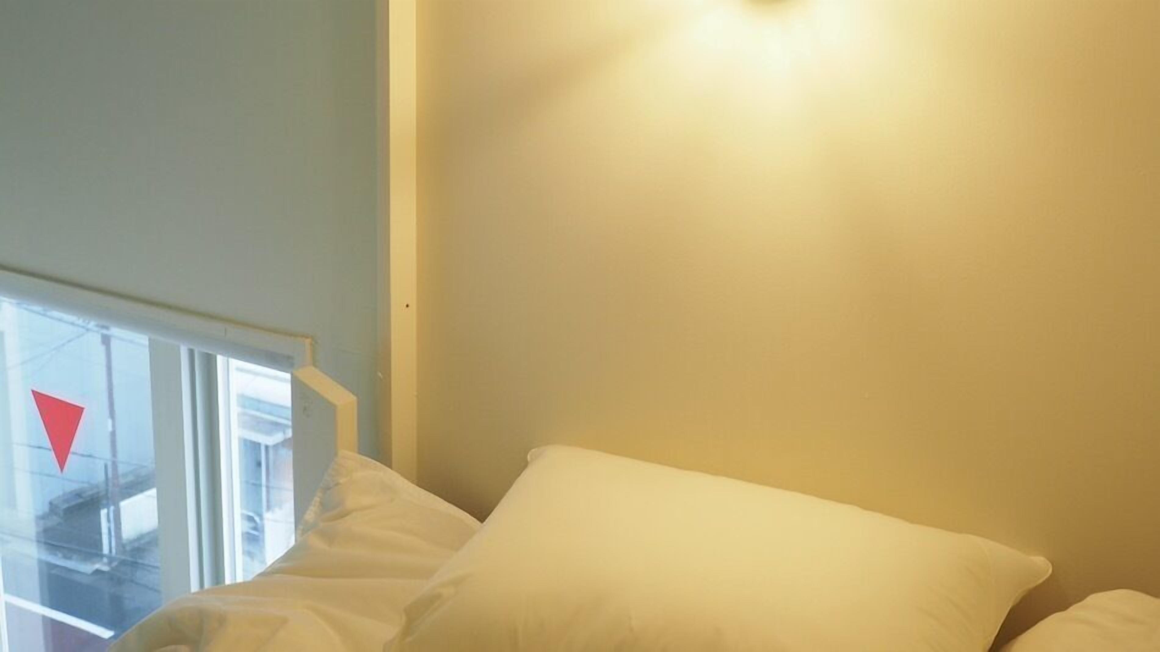 Une pièce avec un lit, une fenêtre ornée d’un panneau triangulaire rouge et un luminaire fixé au mur.