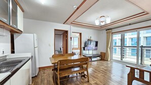 A spacious kitchen with wooden flooring, a dining area with a wooden table and chairs, a built-in TV, and large windows offering a view of the city. - Seongsan Sunrise Pension (Seogwipo)