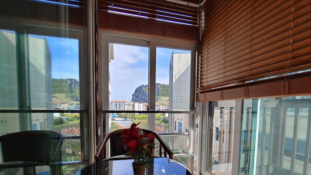 A balcony with a glass table and chairs, a potted plant, and a view of buildings and mountains. - Seongsan Sunrise Pension (Seogwipo)