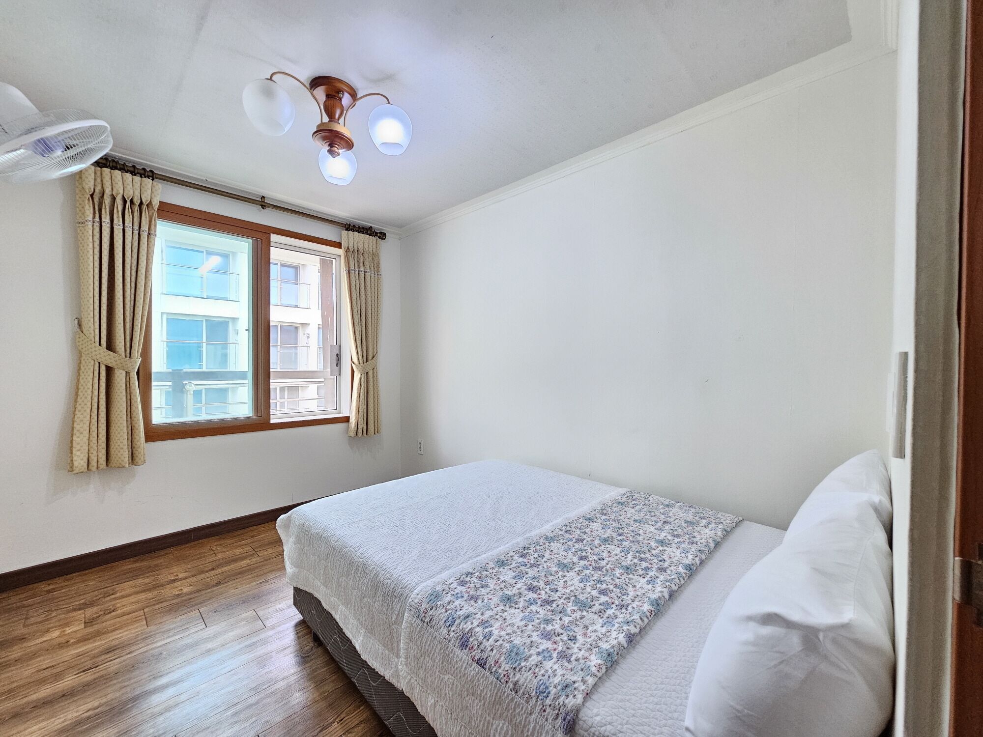 Un dormitorio con cama, ventilador de techo, ventana con cortinas y vistas a edificios.