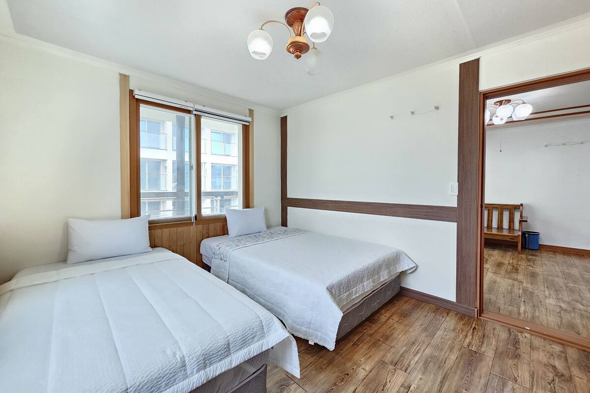Une chambre avec deux lits, un plancher en bois, une fenêtre avec des stores, un luminaire au plafond et un banc en bois dans un coin.