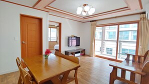 A dining area with a wooden table and chairs, a television on a stand, and a large window with curtains. - Seongsan Sunrise Pension (Seogwipo)