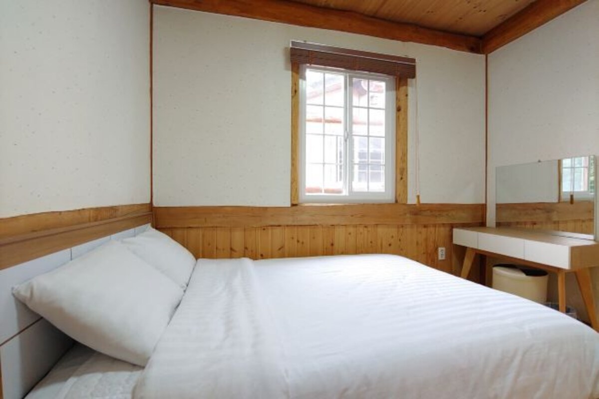 Een slaapkamer met een houten hoofdeinde, een enkel bed, een nachtkastje, een spiegel en een raam met houten kader.