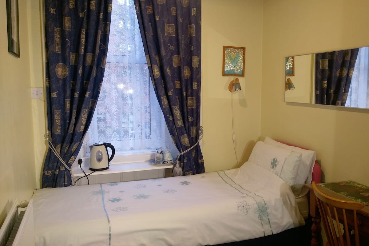 A small, neatly arranged bedroom with a single bed, a bedside table, a mirror, and a window with curtains.