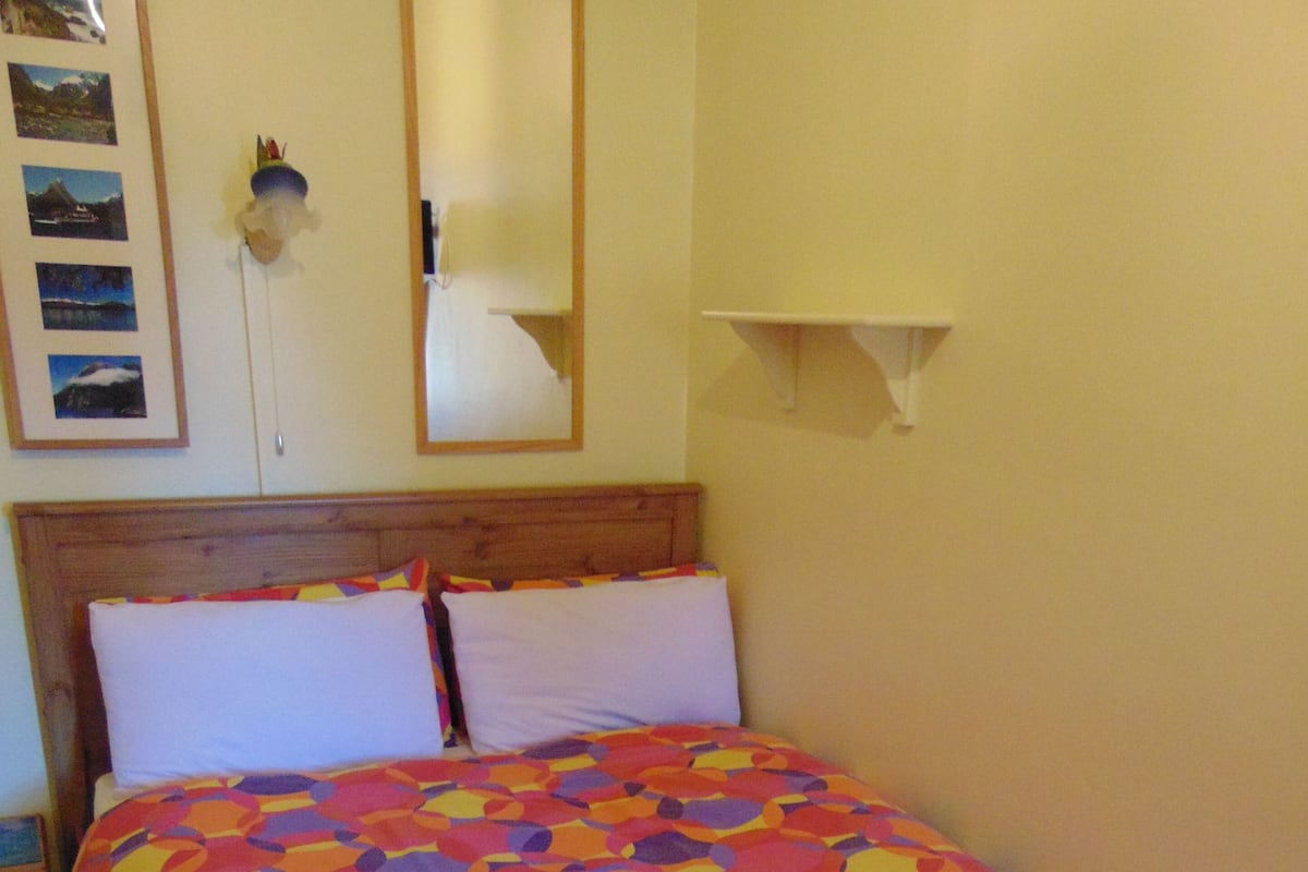 A bed with a colorful geometric-patterned bedspread, a wooden headboard, and a wall-mounted shelf with a mirror.