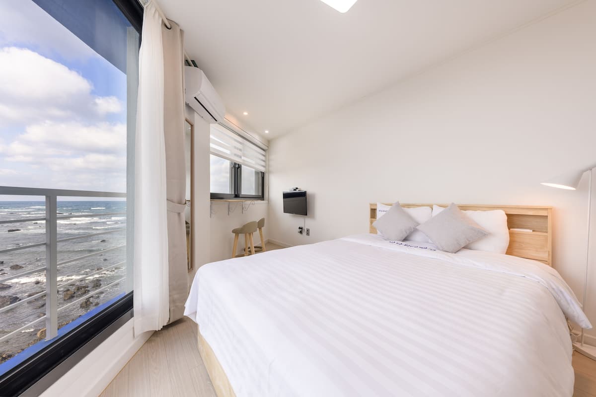 A bedroom with a large bed, a TV mounted on the wall, and a view of the beach through a large window.