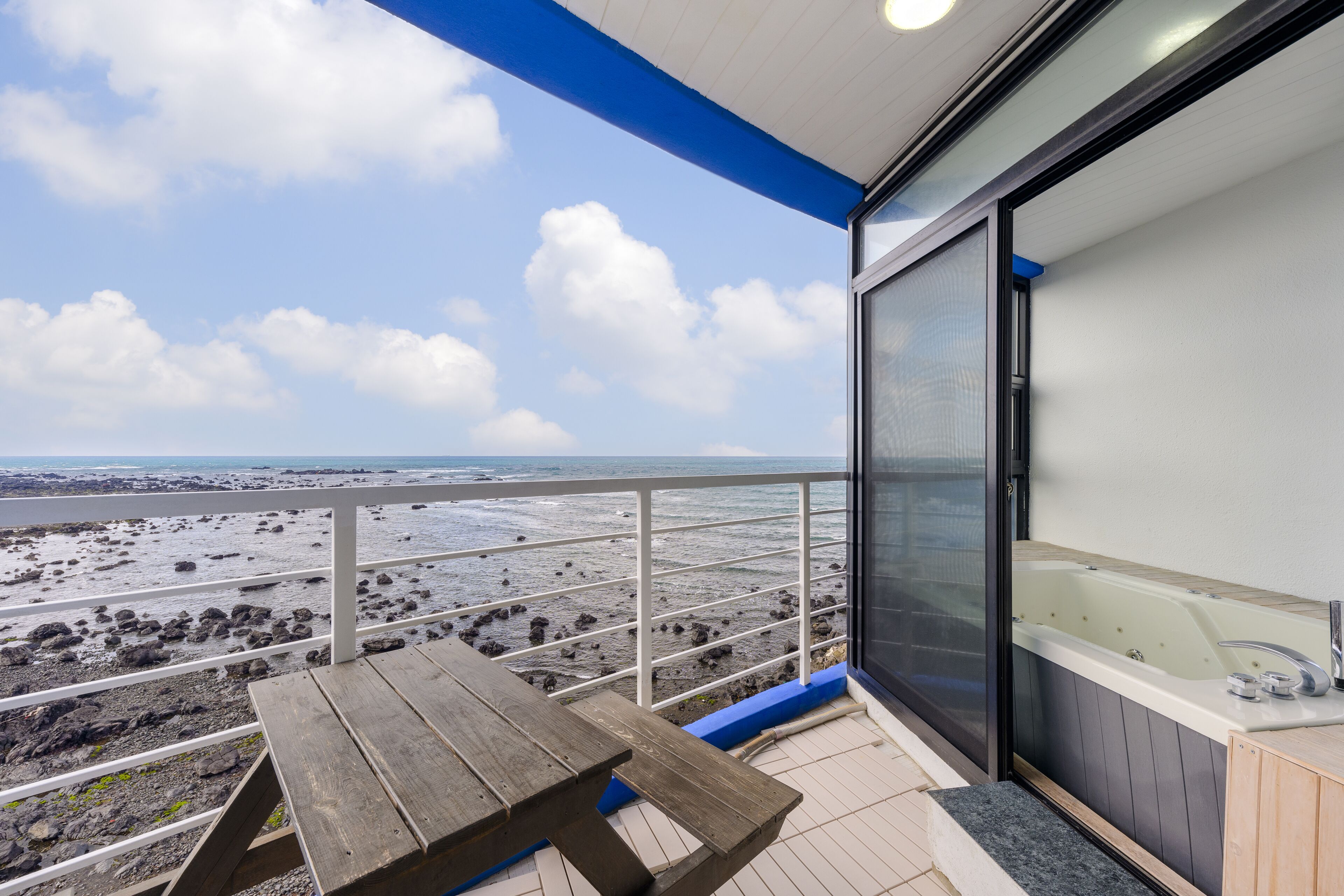 A balcony with a wooden table and a hot tub. The balcony overlooks a rocky beach and the ocean.