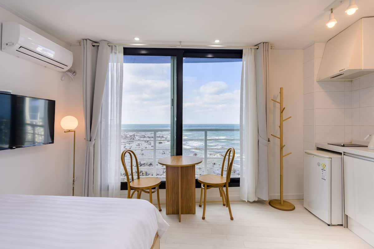 A hotel room with a bed, a TV, a dining table with chairs, a view of the beach, and a kitchenette.