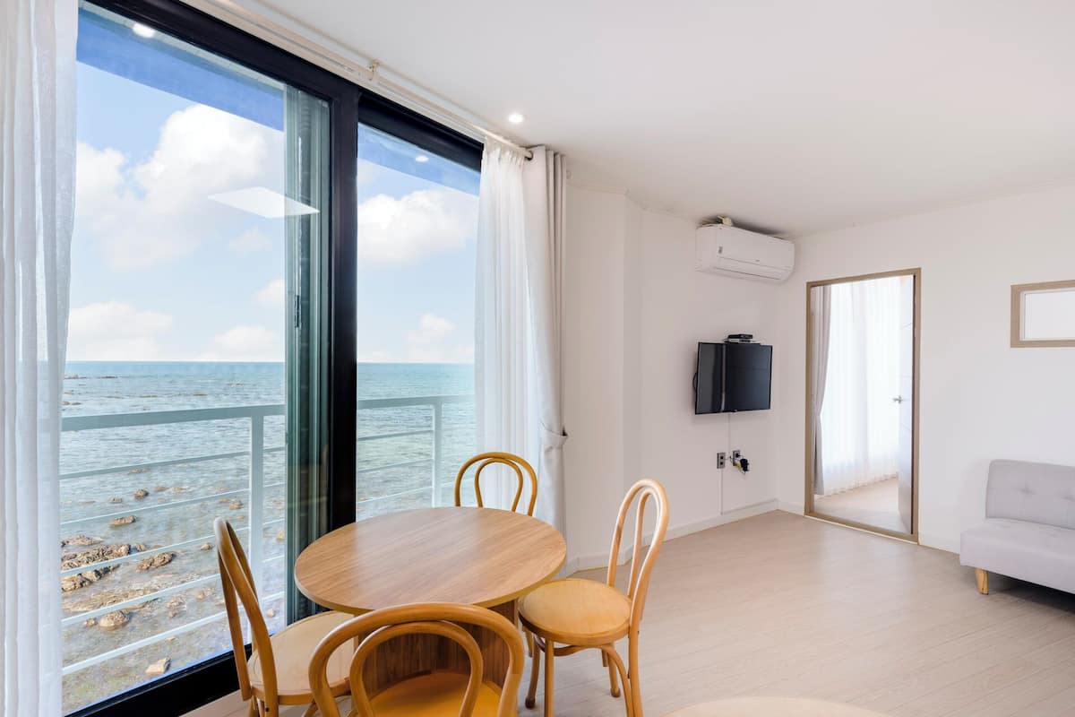 A room with a large window overlooking the sea, a round wooden table with chairs, a flat-screen TV mounted on the wall, and a light-colored sofa.