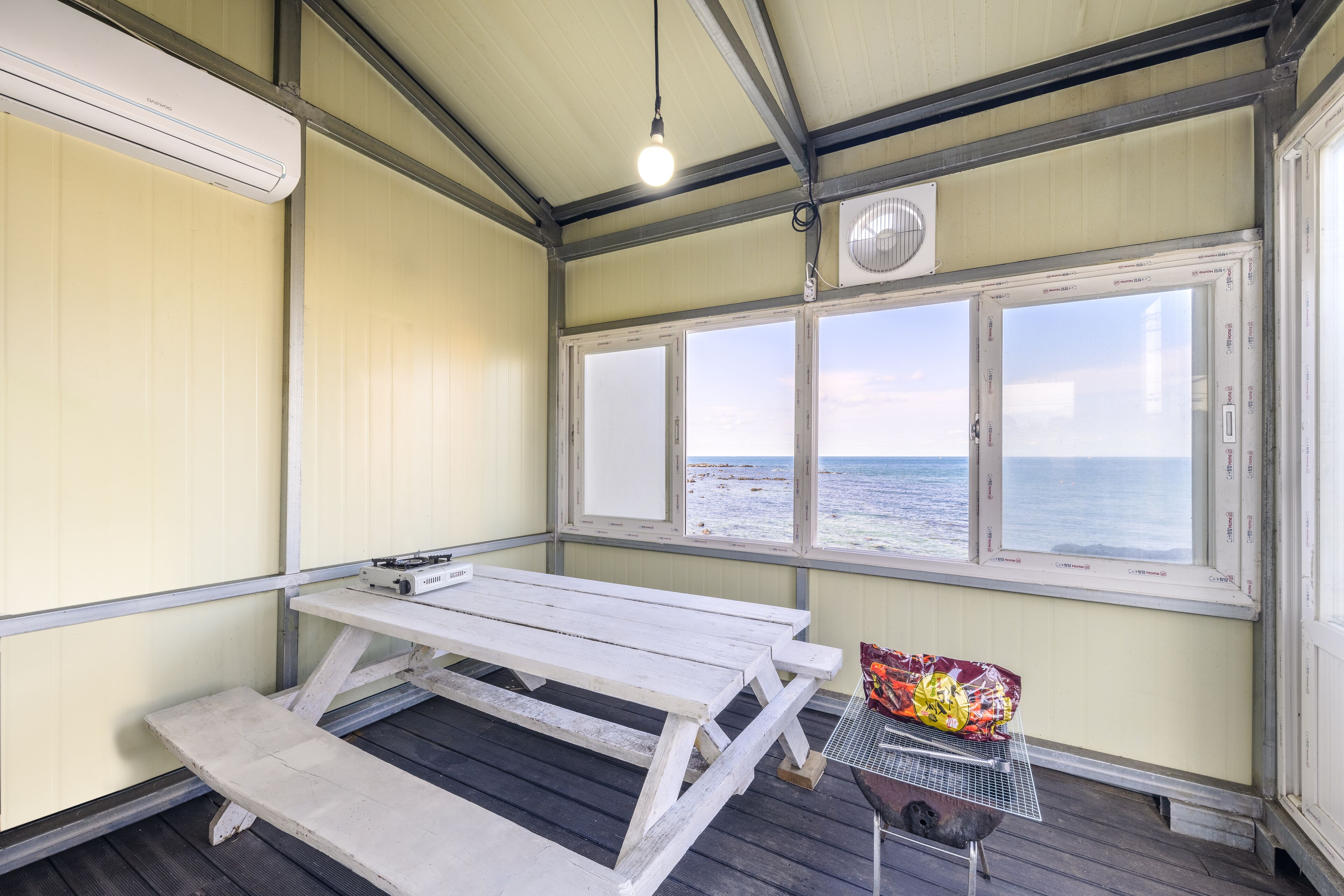 A small, enclosed room with a wooden table and bench, a grill, and a view of the ocean through large windows.