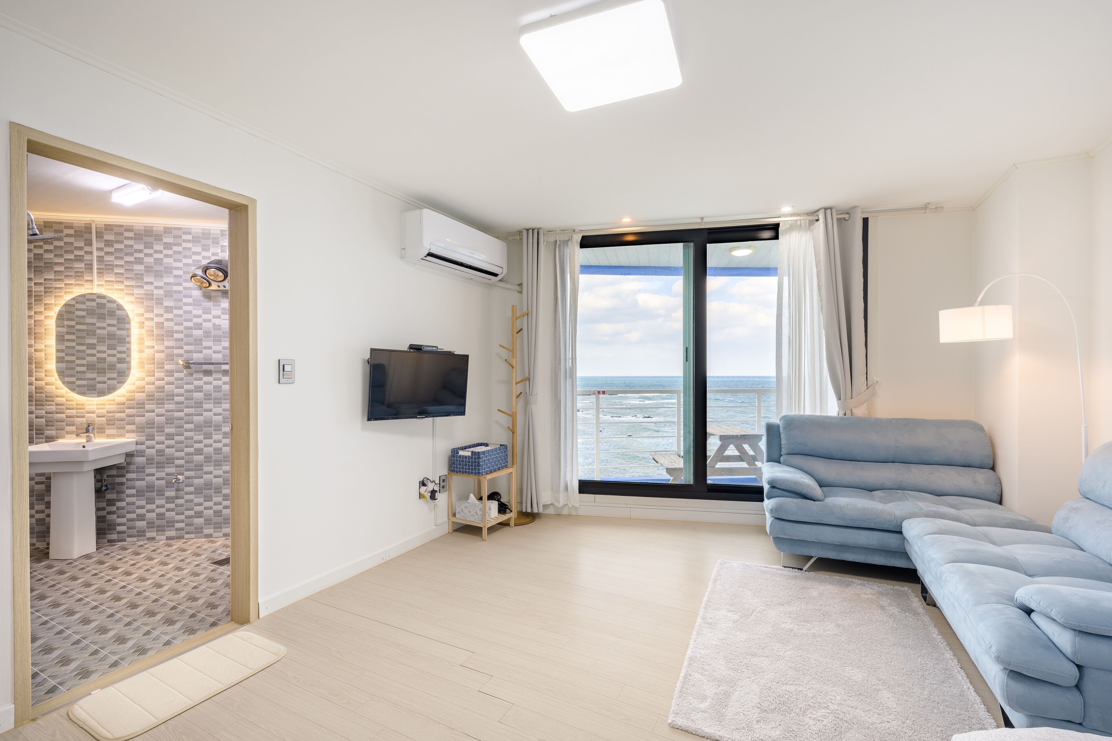 A modern living room with a sofa, a TV, a window with a view of the sea, and a bathroom with a tiled wall and a sink.