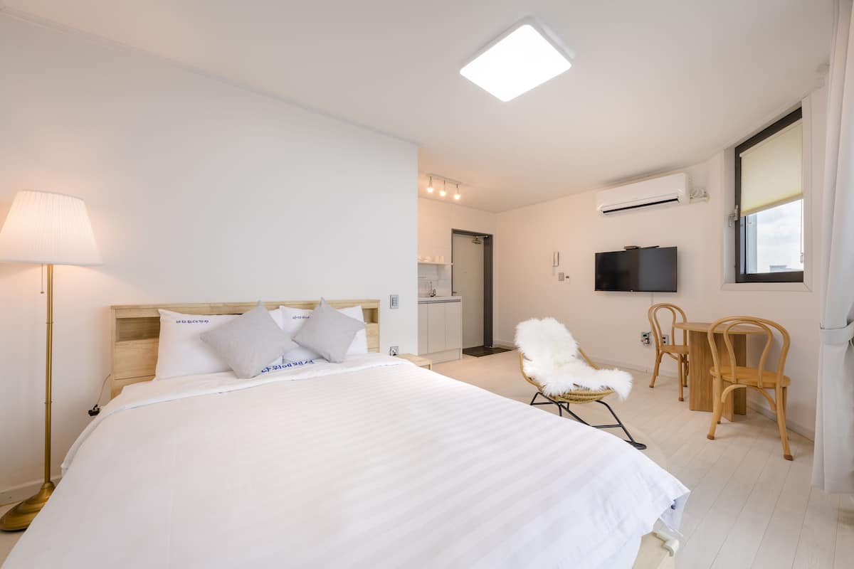 A hotel room with a large bed, a desk, a chair, a TV, and a kitchenette in the background.